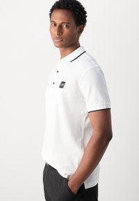 Polo bianco con un tessuto testurizzato, dettagli del colletto neri, placket a tre bottoni e una piccola patch con il logo sul petto.
