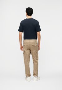 Mann trägt ein marineblaues kurzärmeliges Hemd, beige Cargohosen mit geknöpften Gesäßtaschen und weiße Turnschuhe, steht mit dem Rücken zur Kamera.