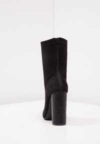 Bota de ante negra hasta la rodilla con cierre de cremallera en la parte posterior y un tacón cuadrado texturizado negro. Superficie suave con un diseño ajustado.