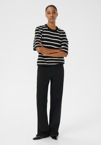 Černobíle pruhovaný pletený svetr s krátkými rukávy, kombinovaný s černými kalhotami s vysokým pasem a širokými nohavicemi a špičatými černými botami.