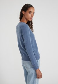 Jeune femme aux cheveux bouclés portant un pull bleu et un jean bleu clair, se tenant de profil contre un fond uni.