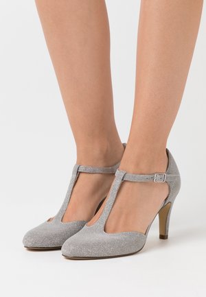 Chaussures à talons hauts argentées avec un design à bride en T, tissu texturé, bout ouvert et un talon fin. Accentué d'une petite boucle réglable.