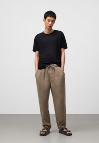 Zwarte lichte t-shirt, beige linnen broek met koord, en zwarte sandalen. De broek heeft een relaxte pasvorm; het t-shirt heeft een ronde hals.