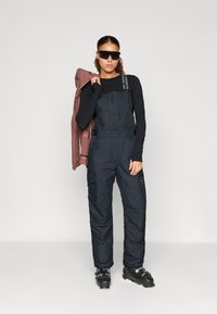 Fekete szigetelt síoverall állítható vállpántokkal, hosszú ujjú fekete toppal és napszemüveggel; egy bordó kabát lóg az egyik karon.