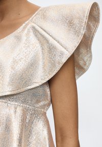 Γυναίκα με αμάνικο φόρεμα με μεταλλική υφή και μεγάλο φουσκωτό λεπτομέρεια στον ώμο, που δείχνει το άνω μέρος του κορμού και το χέρι.