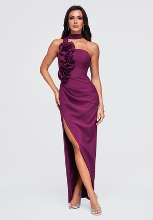 Un mannequin pose sur un fond uni, vêtu d’une robe de soirée prune ornée d’applications florales, avec une encolure haute, une fente jusqu’à la cuisse et des talons scintillants.
