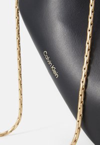Černá kožená kabelka s zlatým řetízkem a embosovaným logem Calvin Klein. Hladký povrch s pevnou strukturou.