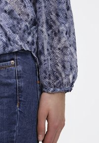 Blouse à manches longues en tissu léger texturé avec un motif serpent bleu et gris ; détail de poignet froncé. Associée à un jean en denim foncé.