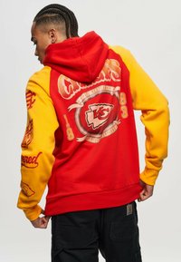 Punainen ja keltainen fleece-huppari, jossa on graafinen logo selässä, joka esittää "1993" sekä joukkueen tietoja. Ribattu hihansuut ja vyötärö.