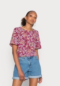 T-shirt de manga curta e decote redondo em cor sangria, com padrões paisley e florais. Usado com calções de ganga azul claro com as bainhas dobradas.