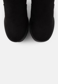 Černé boty na kotník z semiše s kulatou špičkou a hladkou texturou, s prošitým švem kolem podrážky, na bílém pozadí.