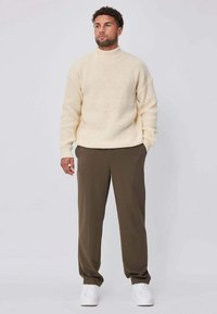 Pull en maille crème avec un col montant, associé à un pantalon vert olive. Le modèle porte des baskets blanches, debout devant un fond uni.