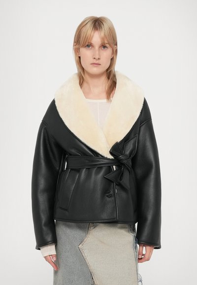 Giacca di pelle nera con un ampio colletto in pelliccia ecologica color crema, vita cinture e maniche oversize. Dettagliata con un fiocco laterale e chiusura a bottone.