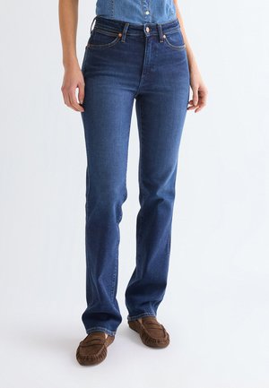 Femeie purtând blugi drepti bleumarin cu talie înaltă și pantofi mocasini maro pe un fundal alb simplu.