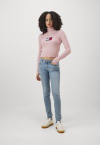 Maglione rosa a coste con collo alto, di lunghezza cropped, con un'etichetta logo. Abbinato a jeans skinny blu chiaro e sneakers bianche con accenti gialli.