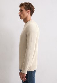 Cremfarbener, strukturiert gestrickter Pullover mit rundem Ausschnitt und langen Ärmeln, kombiniert mit blauer Jeans. Mit gerippten Bündchen und Saum.