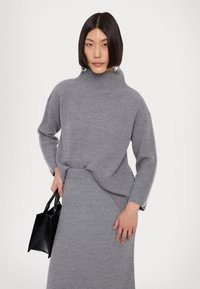 Grå strikket højhalset sweater med ribbet struktur og sidesyninger, parret med en matchende nederdel. Modellen holder en sort håndtaske.