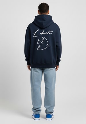 Felpa blu marina con una grande grafica bianca di una colomba e il testo "Liberté" sulla schiena. Abbinata a jeans azzurri e sneakers blu.