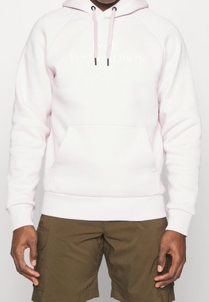 Mand iført lyserød hættetrøje med frontlomme og snore, kombineret med olivengrønne cargoshorts, stående foran en ensfarvet baggrund.