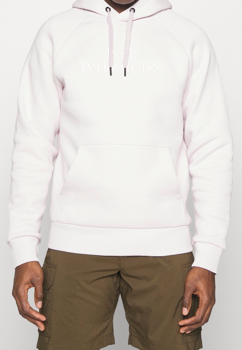 Homme portant un sweat à capuche rose pâle avec une poche avant et des cordons, associé à un short cargo vert olive, debout devant un fond uni.