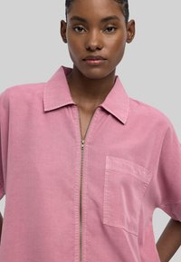 Różowa koszula zapinana na zamek błyskawiczny z krótkimi rękawami, wyprofilowanym kołnierzykiem i kieszonką na piersi. Wykonana z miękkiego materiału o gładkiej fakturze.