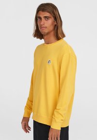 Sudadera de algodón amarilla con cuello redondo, mangas largas y un pequeño logo circular en el pecho. Tela suave y texturizada, corte relajado.