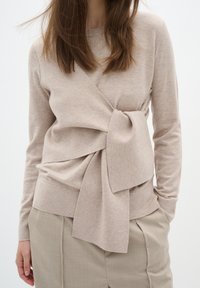 Beige gestrickter Pullover mit Wickeldesign und Schleifendetail in der Taille, kombiniert mit hellbeigen, maßgeschneiderten Hosen. Glatte, weiche Textur.