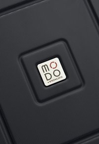 Section de valise texturée noire avec une étiquette de logo carrée indiquant « MODO by RONCATO » au centre.