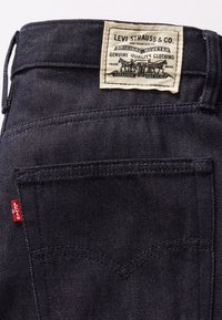 Σκούρο τζην παντελόνι με δερμάτινη ετικέτα στο πίσω μέρος, που διαθέτει ανάγλυφο κείμενο και κόκκινη ετικέτα Levi’s. Περιλαμβάνει διπλή ραφή και πίσω τσέπες.