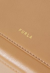 Tan leren oppervlak met een gestructureerde afwerking, voorzien van een goudgeolvde "FURLA"-logo en nette stiksels langs de randen.