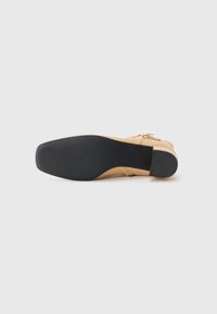 Beige hak schoen met een zwarte rubberen zool, met een glad leren bovenkant en een goudkleurige gesp accent aan de zijkant.
