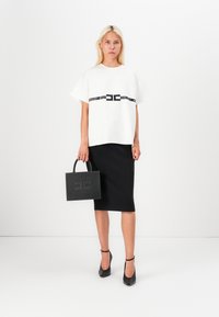 White short-sleeve top with a black band detail, paired with a black pencil skirt. Model holds a black textured handbag with embossed logo.