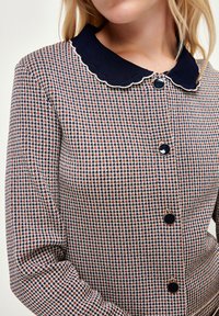 Kvinna som bär en texturerad långärmad jacka med marinblå vågig krage och stora svarta knappar fram.