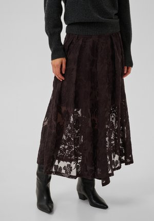Femme portant une jupe midi en dentelle florale foncée, des bottines noires à bouts pointus et un pull gris foncé à manches longues, debout sur un fond uni.