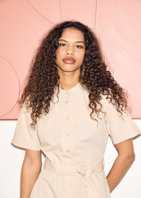 Vrouw met lang krullend haar, gekleed in een beige jurk met knopen en een gestrikte taille, staat tegen een perzikkleurige abstracte achtergrond.