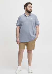 Polohemd in Heatherblau mit weißem Kragen, kombiniert mit khakifarbenen Shorts. Das Modell trägt weiße Sneaker. Schlichtes Design, legere Passform.