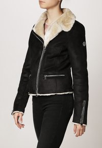 Svart faux mockajacka med en krämfärgad faux pälskrage, dragkedjefickor och silverfärgade detaljer. Har en asymmetrisk dragkedjestängning.