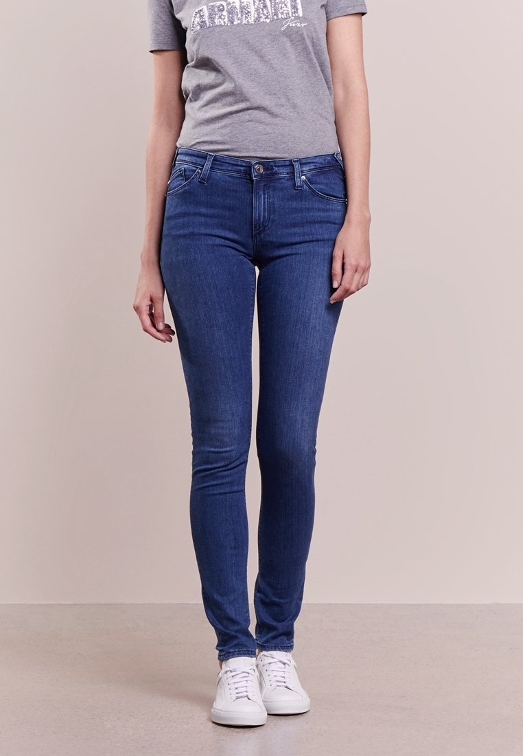 Personne portant un jean skinny bleu, un t-shirt gris avec un texte, et des baskets blanches, debout devant un fond uni.