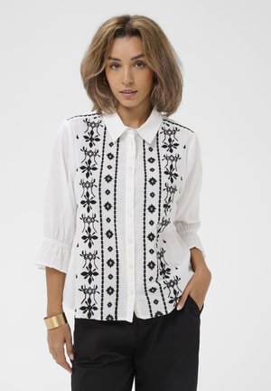 Femeie purtând o bluză albă cu nasturi, cu broderie florală verticală neagră, pantaloni negri și o brățară lată de aur la încheietura mâinii stângi.