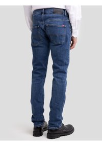 Kék farmernadrágot és fekete csizmát viselő férfi, deréktól lefelé látható fehér ing, egyszínű háttér előtt áll.