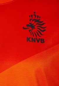 Pomarańczowy sportowy strój z teksturowanym wzorem, z czarnym logo lwa i napisem "KNVB" po lewej stronie.