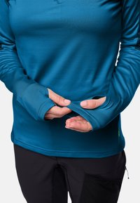 Maglietta a maniche lunghe di colore teal con fori per i pollici, realizzata in tessuto liscio. Presenta un collo a giro e cuciture a contrasto. Indossata con pantaloni scuri.