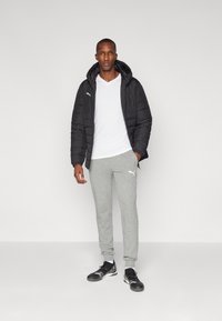 Sort dunjakke med hætte, hvid v-hals t-shirt, grå sweatpants med logo og sorte træningssko. Enkel, funktionel sportstøj.
