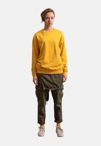 Felpa gialla a maniche lunghe, pantaloni cargo oliva con cerniere e tasche frontali, sneakers beige e calzini gialli. Design semplice, vestibilità casual.