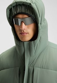 Grüne, gepolsterte Jacke mit hohem Kragen und übergroßer Kapuze. Begleitet von verspiegelten Sonnenbrillen, die eine glatte, matte Textur zeigen.