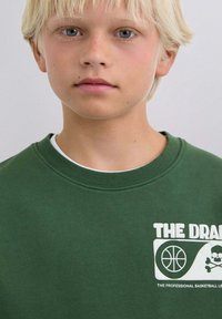 Sudadera verde con un gráfico blanco que muestra un diseño de baloncesto y calavera. Cuello redondo acanalado y tejido texturizado.