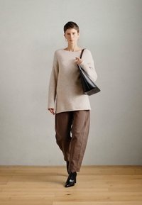 Osoba s krátkymi vlasmi, oblečená v béžovom svetri, hnedých nohaviciach a čiernych členkových čižmách, nesúca čiernu kabelku na pleci.