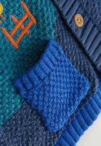 Pleteni izdelek z teksturami v barvi zeleno-modre in mornarsko modre, z modrim žebljičastim žepom ter oranžnim vezenim detajlom. Vidna je tudi okrasna lesena gumb.