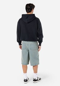 Svart hettegenser med stor hette, kombinert med lyseblå denimshorts som har baklommer og synlige sømmer. Brukt med hvite sokker og mørke joggesko.