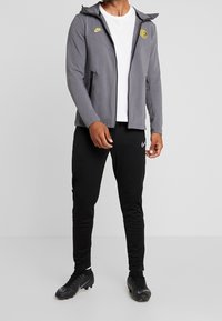 Veste zippée grise Nike avec capuche, poches zippées noires, et accents jaunes, portée avec un pantalon de sport noir et des chaussures de football noires.